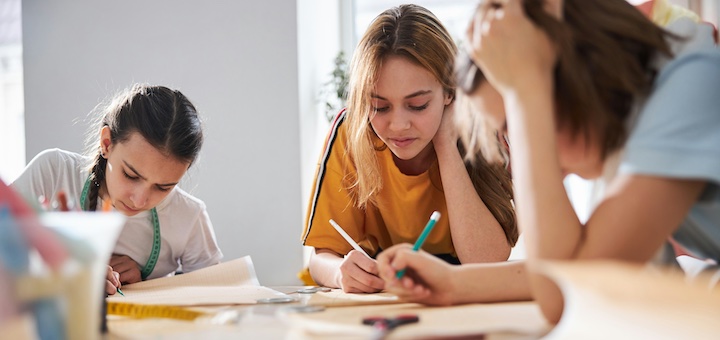 Three girls study together at a bright table, focused on writing in notebooks and papers in a classroom setting.